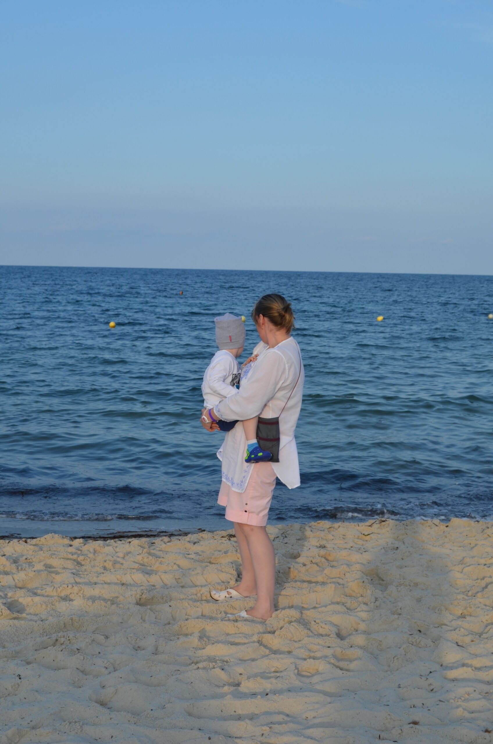 A woman holding her baby at the seaside during a warm summer evening, creating a serene family moment.