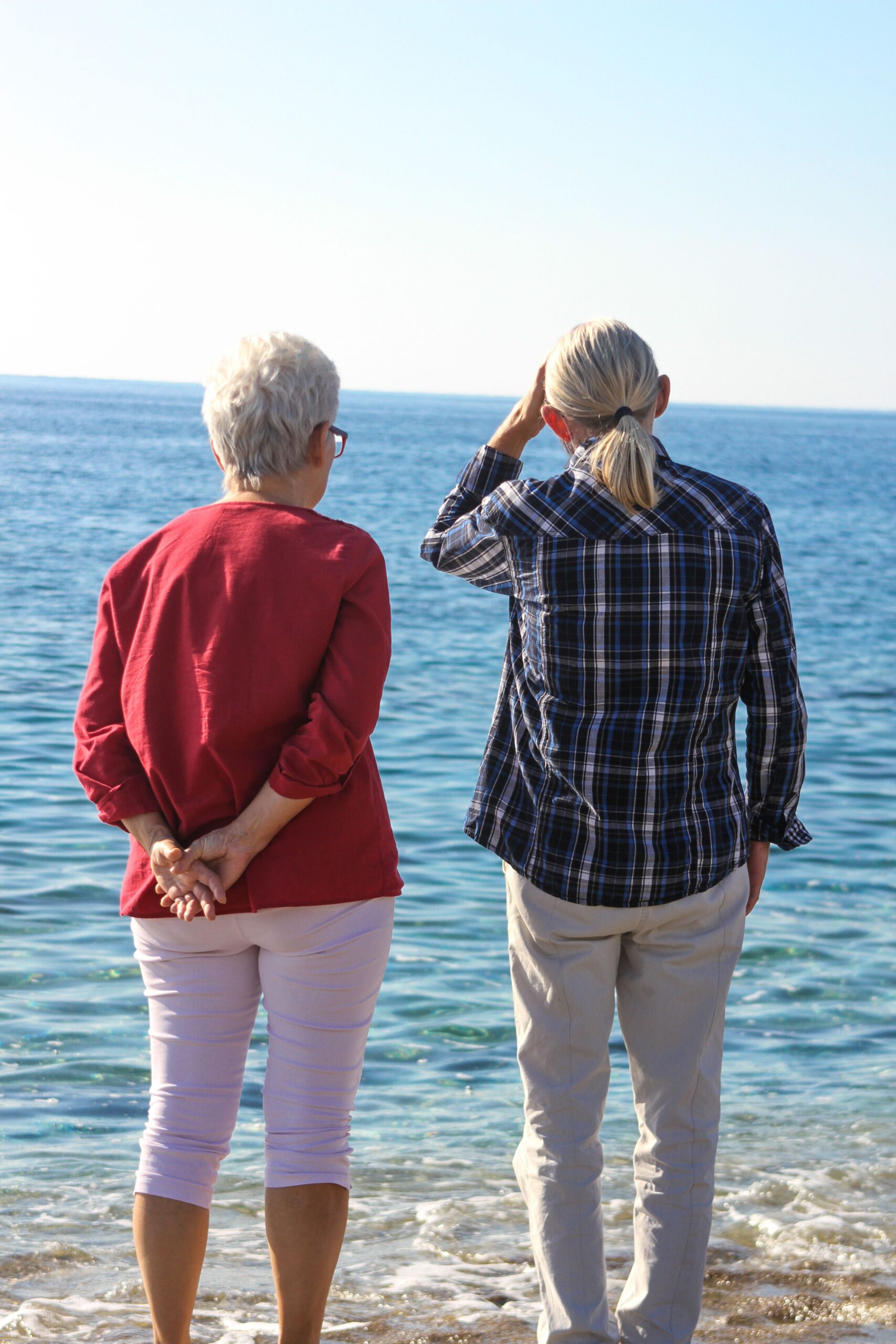 Two senior women stand on a serene beach, gazing at the calm sea under the clear blue sky.